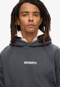 Sudadera con capucha en gris oscuro con el logo blanco "SUPERDRY". Presenta una textura suave, bolsillo tipo canguro y capucha ajustable con cordón.
