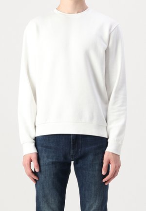 Person iført en ensfarvet hvid sweatshirt med lange ærmer med ribstrikkede manchetter og kant, kombineret med mørkeblå denimjeans, mod en ensfarvet lys baggrund.