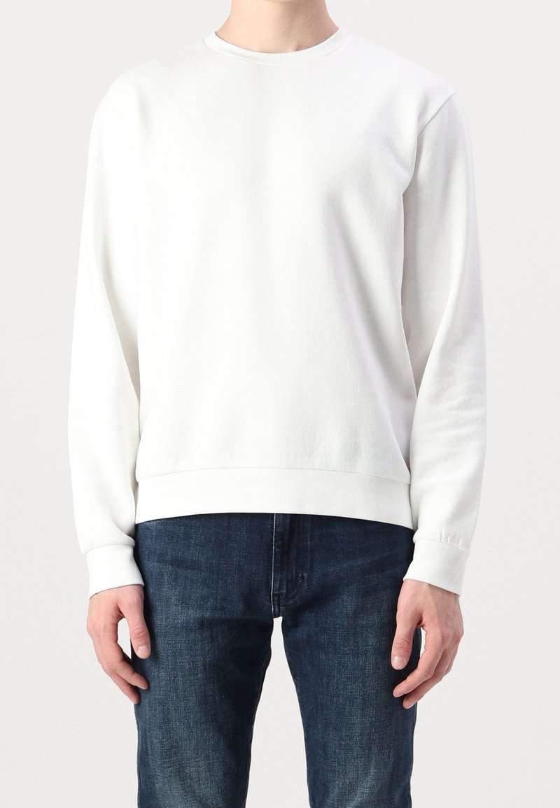 Personne portant un sweat-shirt blanc uni à manches longues avec poignets et ourlet côtelés, associé à un jean en denim bleu foncé, sur un fond clair uni.