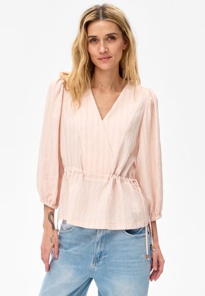 Kobieta o blond włosach ma na sobie jasnoróżową bluzkę z kopertowym dekoltem i rękawami do łokcia oraz jasnoniebieskie dżinsy, pozując na jednolitym tle.