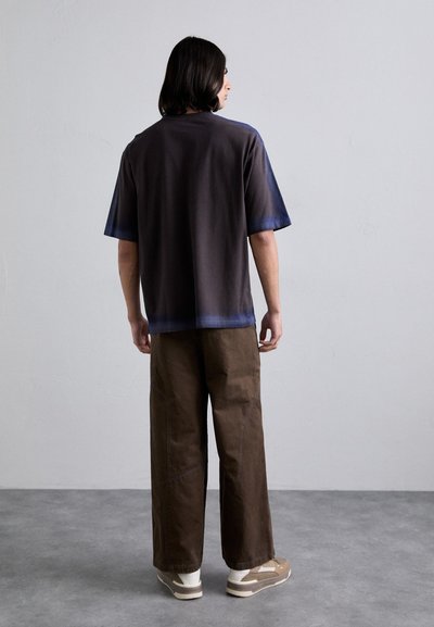 Short-sleeve, gradient T-shirt in dark gray to blue, paired with loose brown pants. Smooth fabric and relaxed fit. White and beige sneakers.