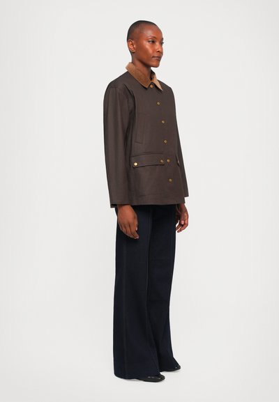 Person wearing a dark brown buttoned jacket with a tan collar and navy wide-leg pants, standing against a plain white background.