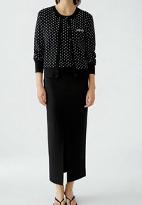 Svart cardigan med vita prickar, knäppt fram, tillsammans med en svart knälång kjol med en sidoklyfta.