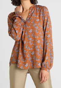 Rustkleurige blouse met een bloemenpatroon in paars en groen, met een v-hals en lange mouwen met gerimpelde manchet.