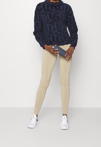 Marinblå hooded sweatshirt med texturerat logomönster, tillsammans med beige leggings och vita träningsskor, håller en grå vattenflaska.