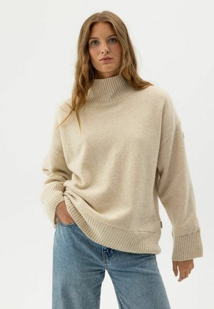 Jauna sieviete, kas valkā bēša krāsas ribotu augstu apkakli un zilas džinsu bikses, stāv ar vienu roku kabatā vienkārša fona priekšā.