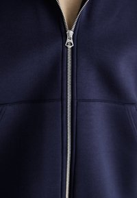 Veste à zip bleu marine en tissu doux, dotée d'une fermeture éclair en métal et de poches avant. Texture lisse et design minimaliste.