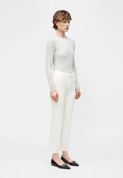 Γυναίκα που στέκεται, φοράει ανοιχτό γκρι μακρυμάνικη εφαρμοστή μπλούζα, λευκό παντελόνι με ίσια γραμμή και ψηλή μέση, και μαύρες μυτερές μπαλαρίνες, σε απλό φόντο.