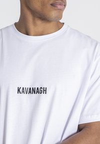 Camiseta de algodón blanca de manga corta. Presenta la palabra "KAVANAGH" en letras mayúsculas negras en el frente. Diseño simple y sin adornos.