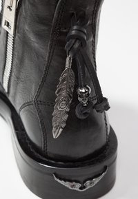 Botín de cuero negro con cremallera plateada, cordones decorativos de cuero negro, charms de metal en forma de pluma y calavera, y un acento redondo en el tacón.