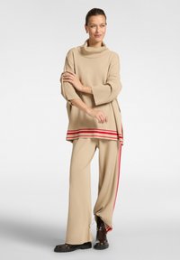Beige coltrui met rode streepaccenten, gecombineerd met bijpassende beige hoge broek met zijden trekkoorddetails en zwarte laarzen.