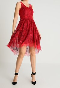 Femme portant une robe rouge en dentelle sans manches avec un ourlet asymétrique et des talons noirs à bride autour de la cheville, debout devant un fond clair uni.