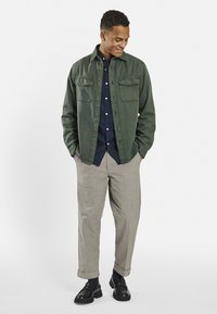 Green cotton shirt jacket with two chest pockets, paired with a navy button-up shirt, gray trousers, and black shoes. Smooth texture.