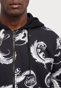 Čierna mikina s kapucňou na zips z bavlny s bielym vírivým abstraktným vzorom. Obsahuje kapucňu so šnúrkou a strieborné detaily zipsu.