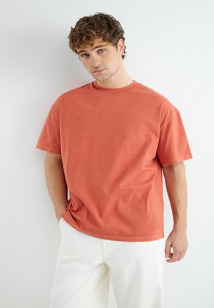 Junger Mann mit lockigem braunem Haar, der ein locker sitzendes oranges T-Shirt und weiße Hosen trägt, steht vor einer schlichten weißen Wand.