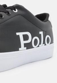 Černé tenisky s bílým logem "Polo", hladký syntetický materiál, kulatá špička a bílá gumová podrážka. Otvory pro šněrování s černými tkaničkami.