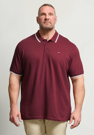 Hombre de constitución musculosa que lleva una camiseta polo burdeos con ribetes blancos y pantalones beige, de pie frente a un fondo claro y liso.