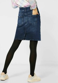 Falda de mezclilla en azul oscuro con corte recto, bolsillos traseros y una abertura en el dobladillo. Lleva puestas medias negras y zapatillas claras.