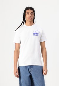 Hvit bomulls T-skjorte med rund hals, med et blått grafisk trykk på venstre bryst som leser "OBEY NEW VISIONS." Brukt sammen med blå jeans.