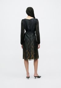 Schwarzes Kleid mit transparenten, langen Ärmeln, einem Gürtel in der Taille und einem goldenen Schlangenprint-Muster durchgehend. Midilanges Design mit figurbetonter Silhouette.