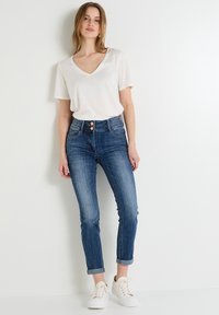 Camiseta blanca de manga corta y escote en V combinada con jeans de mezclilla azul de tiro alto con cierre de botones y tobillos ajustados, usados con zapatillas blancas.