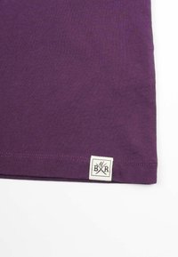 Lila bomull t-shirt med en sydd fåll och en fyrkantig etikett som visar bokstäverna "B" och "R" korsade, vilket indikerar varumärkesidentifikation.