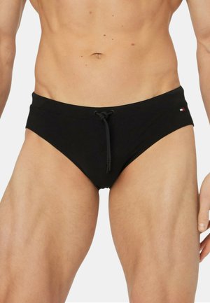 Torace maschile con slip da bagno nero con coulisse frontale e piccolo logo sull'anca destra su sfondo uniforme.
