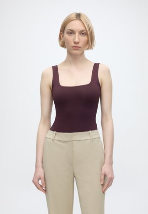 Kvinde med kort blondt hår iført gyldne hoop-øreringe, en tætsiddende ærmeløs mørkelilla top og beige bukser med høj talje, stående frontalt.