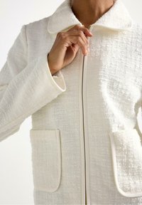 Giacca bianca testurizzata con collo, zip frontale e due tasche a toppa. Accentuata da un design pulito e minimale.