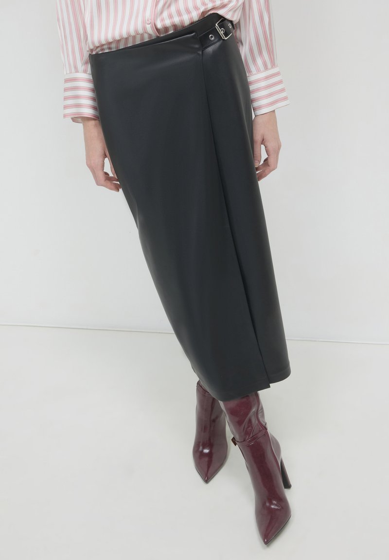 Femme portant une jupe midi en cuir noir avec une boucle sur le côté et des bottes à talons aiguilles bordeaux, assortie à une blouse rose à rayures.