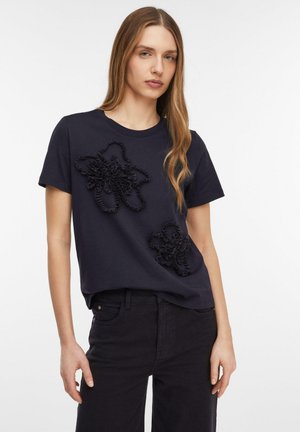 Kobieta o długich włosach, ubrana w granatowy T-shirt z dwoma teksturowanymi kwiatowymi wzorami, w czarnych spodniach, stojąca na białym tle.