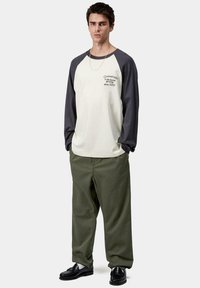 Joven de pie que lleva una camiseta de béisbol de manga larga en crema y negro, pantalones verde olivo, mocasines negros y un collar de cadena de plata.
