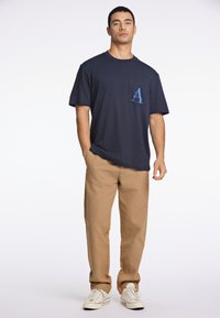 Mörkblå T-shirt med en ficka som har ett ljust blått bokstav "A", parat med beige byxor och vita sneakers med detaljer.