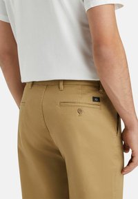 Pantalon en coton couleur beige avec une texture lisse, doté de poches arrière, d'un bouton en accent, et d'une petite étiquette de marque sur la poche arrière droite.