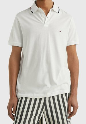 Une personne portant un polo blanc à manches courtes avec un col à rayures et un pantalon à rayures verticales noir et blanc.