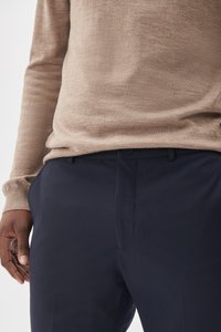 Pull en tricot marron clair associé à un pantalon en toile bleu marine. Le pull présente une texture subtile, tandis que le pantalon a une finition lisse et une fermeture à bouton.