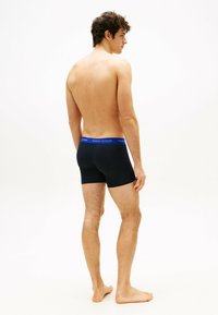 Boxer-briefs noirs avec une ceinture de marque bleue, fabriqués à partir d'un matériau extensible. Le modèle se tient pieds nus, légèrement de côté, montrant l'ajustement et la coupe.