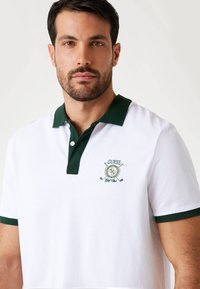 Weißes Poloshirt mit dunkelgrünem Kragen und Bündchen. Mit gesticktem "GUESS Golf Club"-Logo und golfbezogenen Grafiken.