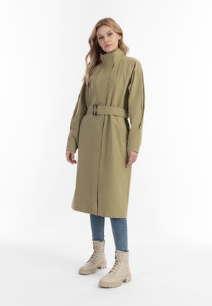 Vrouw in een olijfgroene trenchcoat met riem, lichte spijkerbroek en beige veterschoenen, staand met een neutrale uitdrukking op een witte achtergrond.