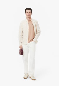 Randig kräm- och beige skjorta med knappar över en ljusrosa polo, matchat med vita byxor och krämfärgade sneakers. Håller en liten vinröd väska.