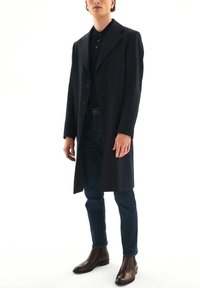 Palton din lână bleumarin cu rever ascuțit și cinci nasturi pe față, purtat peste un tricou polo negru și blugi albaștri, asortat cu cizme din piele maro până la gleznă.
