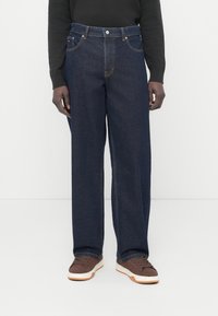 Σκούρο μπλε τζιν με φαρδιά μπατζάκια από denim με παραδοσιακό σχεδιασμό πέντε τσεπών και αντίθετη ραφή· φορεμένα με καφέ αθλητικά παπούτσια.