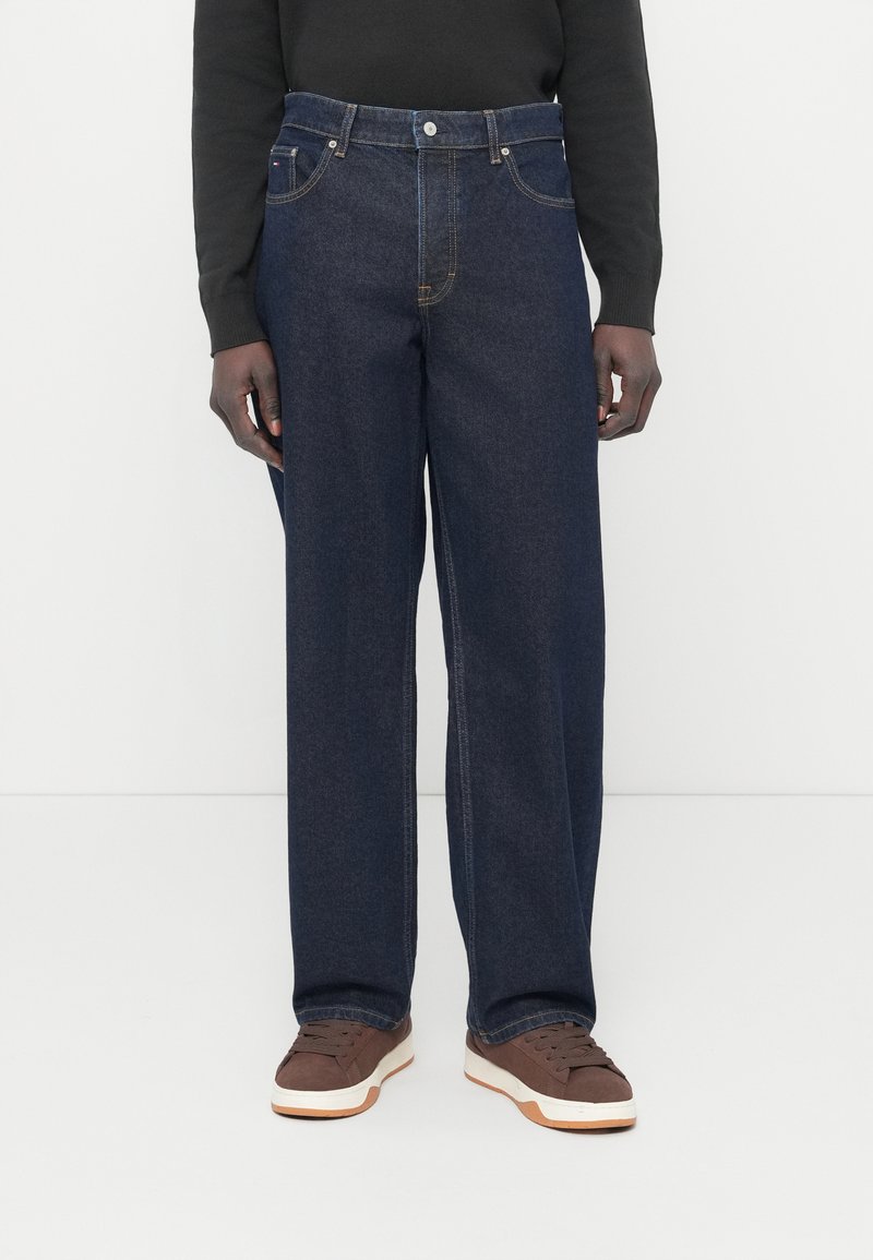 Σκούρο μπλε τζιν με φαρδιά μπατζάκια από denim με παραδοσιακό σχεδιασμό πέντε τσεπών και αντίθετη ραφή· φορεμένα με καφέ αθλητικά παπούτσια.