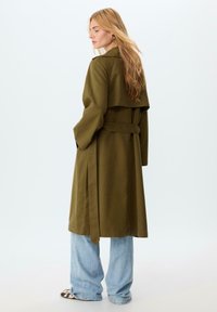 Trench coat verde oliva con vita cinchata, maniche ampie e design posteriore a strati. Indossato sopra jeans a gamba larga di colore blu chiaro e scarpe con fantasia.