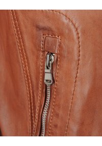 Nabijheidsfoto van een bruinleren jack met een zilveren rits, stiksels en een gladde textuur op een warmgekleurde achtergrond.