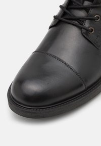 Chaussure en cuir noir à bout arrondi, avec une texture lisse, des accents cousus et une semelle en caoutchouc plate. Les lacets sont noirs, avec des œillets métalliques visibles.
