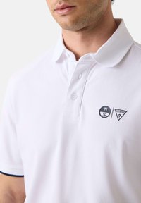 Homme portant un polo blanc avec un petit logo boussole et un logo Guess sur le côté gauche de la poitrine, boutonné au col, sur un fond uni.