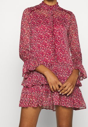 Vrouw draagt een rood bloemenjurk met lange mouwen, ruches aan de kraag, een gelaagde rok en doorzichtig stof, staat met gevouwen handen.