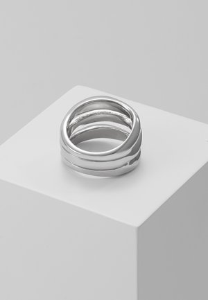 Gestapelde glanzende zilveren ringen met afwisselend gladde en gegrachte banden, gepresenteerd op een witte sokkel tegen een lichtgrijze achtergrond.