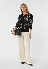Schwarzer Pullover mit cremefarbenem geometrischem Muster, kombiniert mit weißen, weiten Hosen und einer kleinen schwarzen Handtasche, dazu elegante spitze Schuhe.